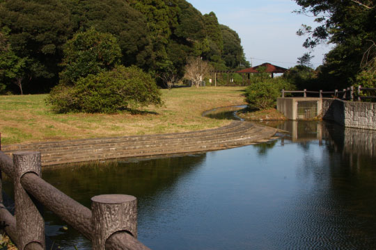 中央に小川が流れ、奥に橋が架かっている公園の写真