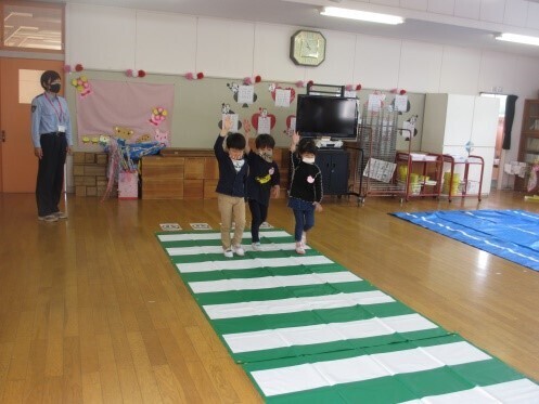 教室の床に敷かれた横断歩道に見立てた緑色と白色のシートの上を歩いている3名の園児の交通安全教室の様子の写真