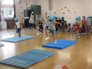 水色の帽子を被った子どもたちが2列に縦に並んでおり、2名の子どもがマットの上を飛び跳ねて移動している様子の写真