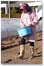 長靴を履き青いバケツを持った女性が畑に肥料を散布している写真