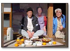 みかんやお茶が置かれた縁側で、鈴木 孝一氏とご家族の方2名がくつろいでいる写真
