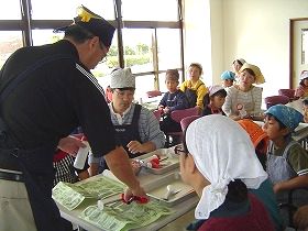 料理講習会の講師が、机の上に置いた用紙を指さししており、参加者の人達が説明を聞いている写真